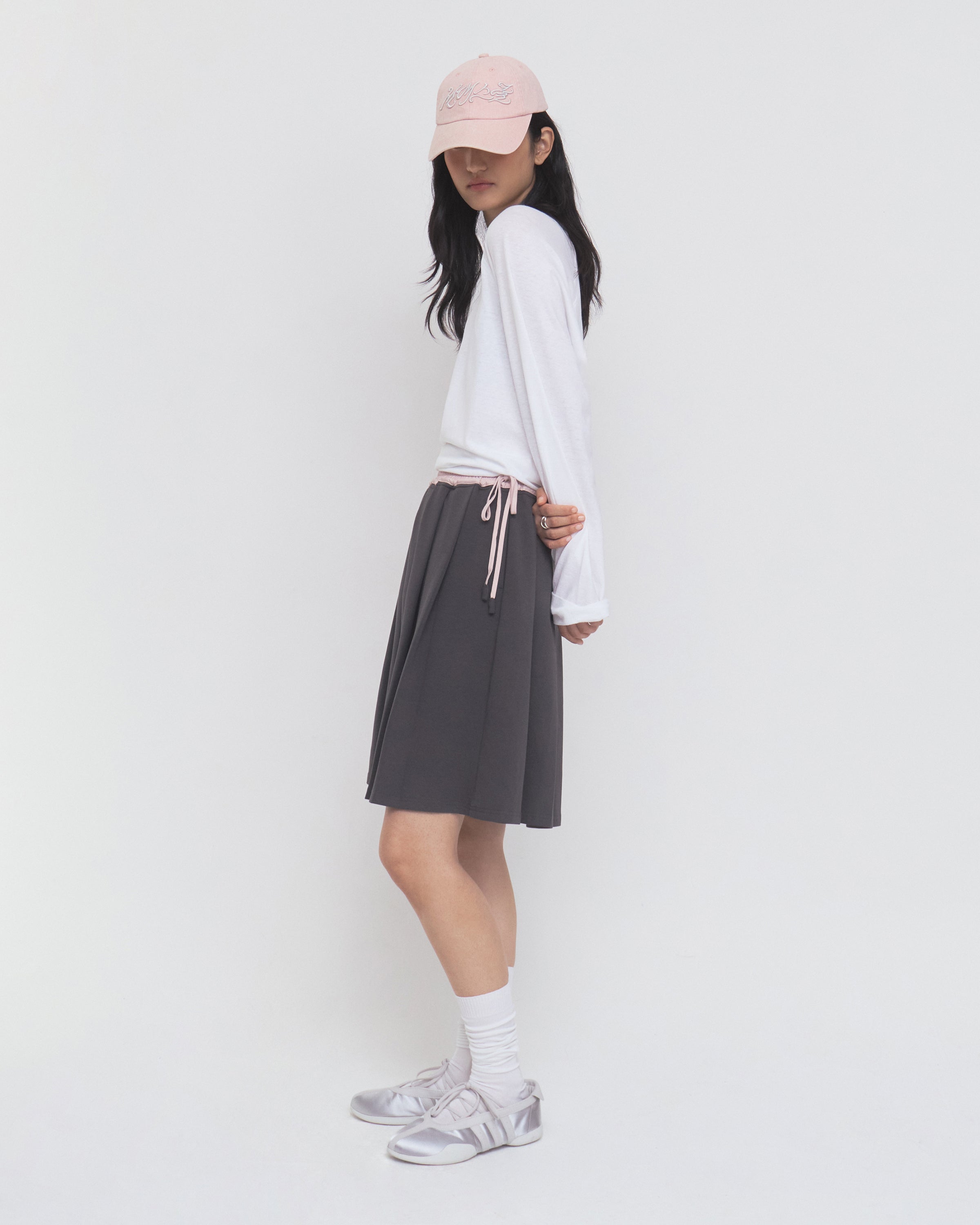 Woman in a long-sleeve white shirt, CHARCOAL KONA MIDI SKIRT by Sundae School, pink cap, white socks, and silver sneakers stands sideways against a plain white background.
