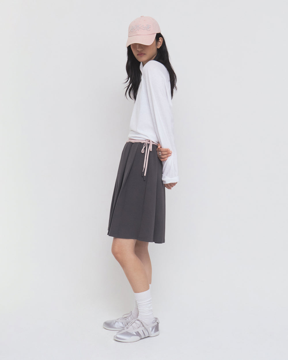 Woman in a long-sleeve white shirt, CHARCOAL KONA MIDI SKIRT by Sundae School, pink cap, white socks, and silver sneakers stands sideways against a plain white background.