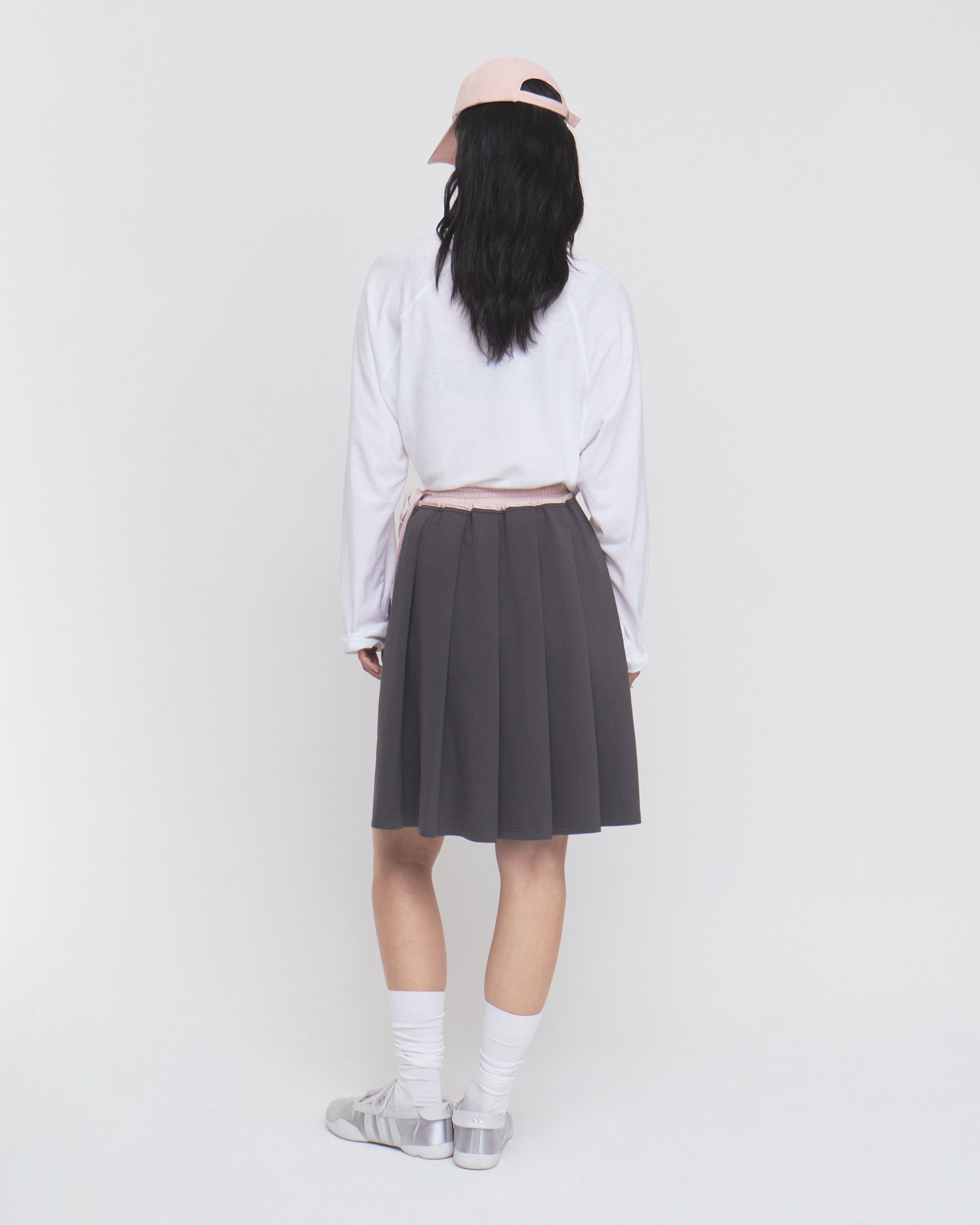 A person with long black hair stands facing away against a plain white background, wearing a light pink cap, white long-sleeve shirt, white socks, gray shoes, and the CHARCOAL KONA MIDI SKIRT by Sundae School.