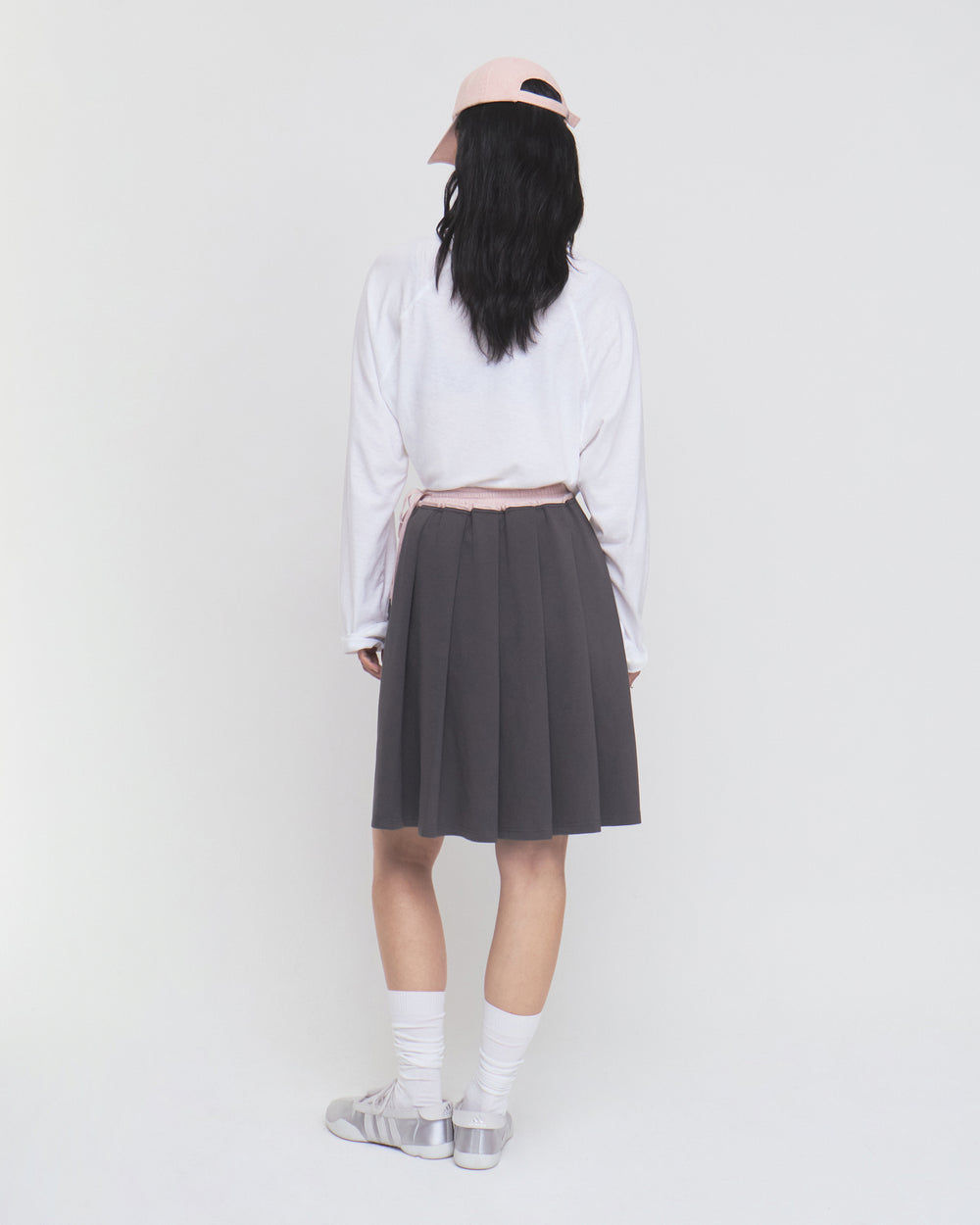 A person with long black hair stands facing away against a plain white background, wearing a light pink cap, white long-sleeve shirt, white socks, gray shoes, and the CHARCOAL KONA MIDI SKIRT by Sundae School.