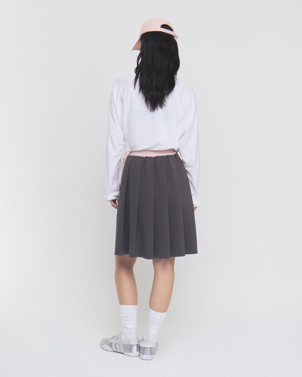 A person with long black hair stands facing away against a plain white background, wearing a light pink cap, white long-sleeve shirt, white socks, gray shoes, and the CHARCOAL KONA MIDI SKIRT by Sundae School.