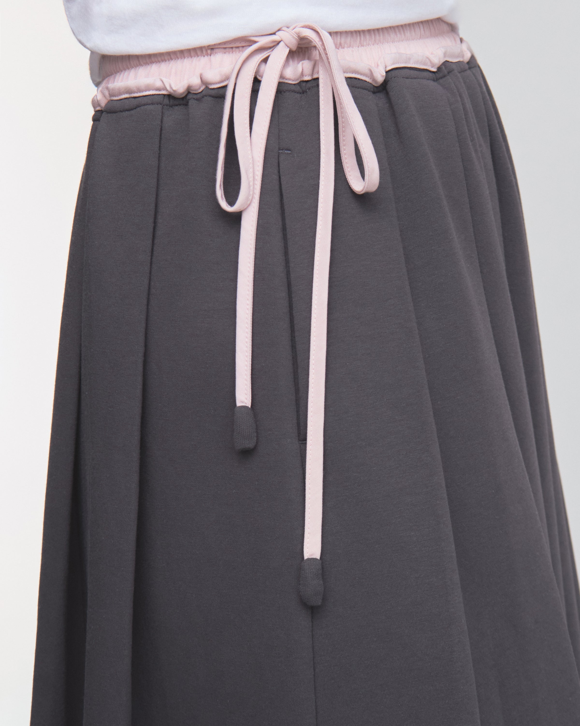 A close-up of a person wearing the CHARCOAL KONA MIDI SKIRT by Sundae School, featuring light pink drawstrings tied in a bow at the waist, paired with a white top.