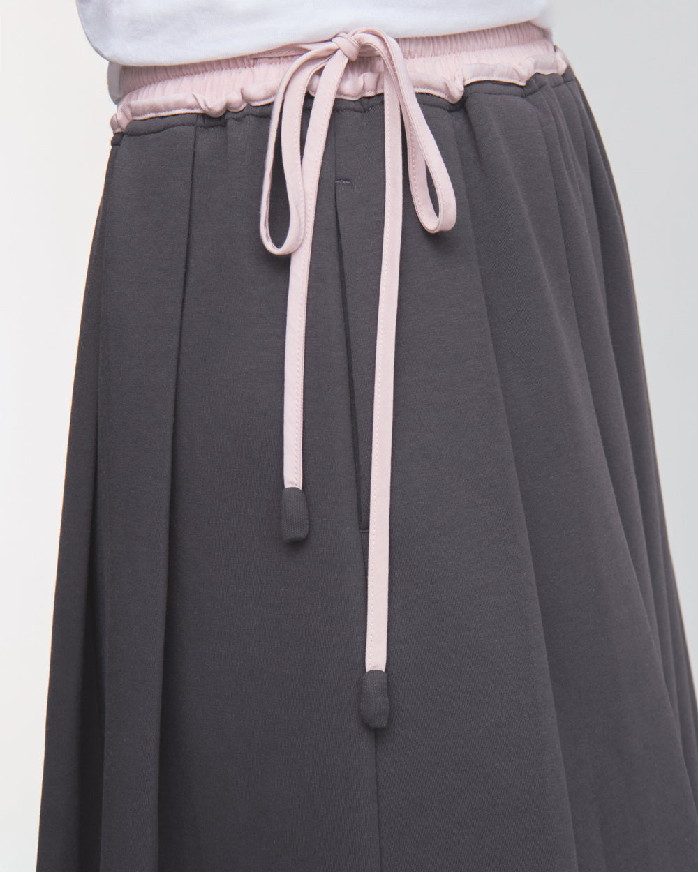 A close-up of a person wearing the CHARCOAL KONA MIDI SKIRT by Sundae School, featuring light pink drawstrings tied in a bow at the waist, paired with a white top.