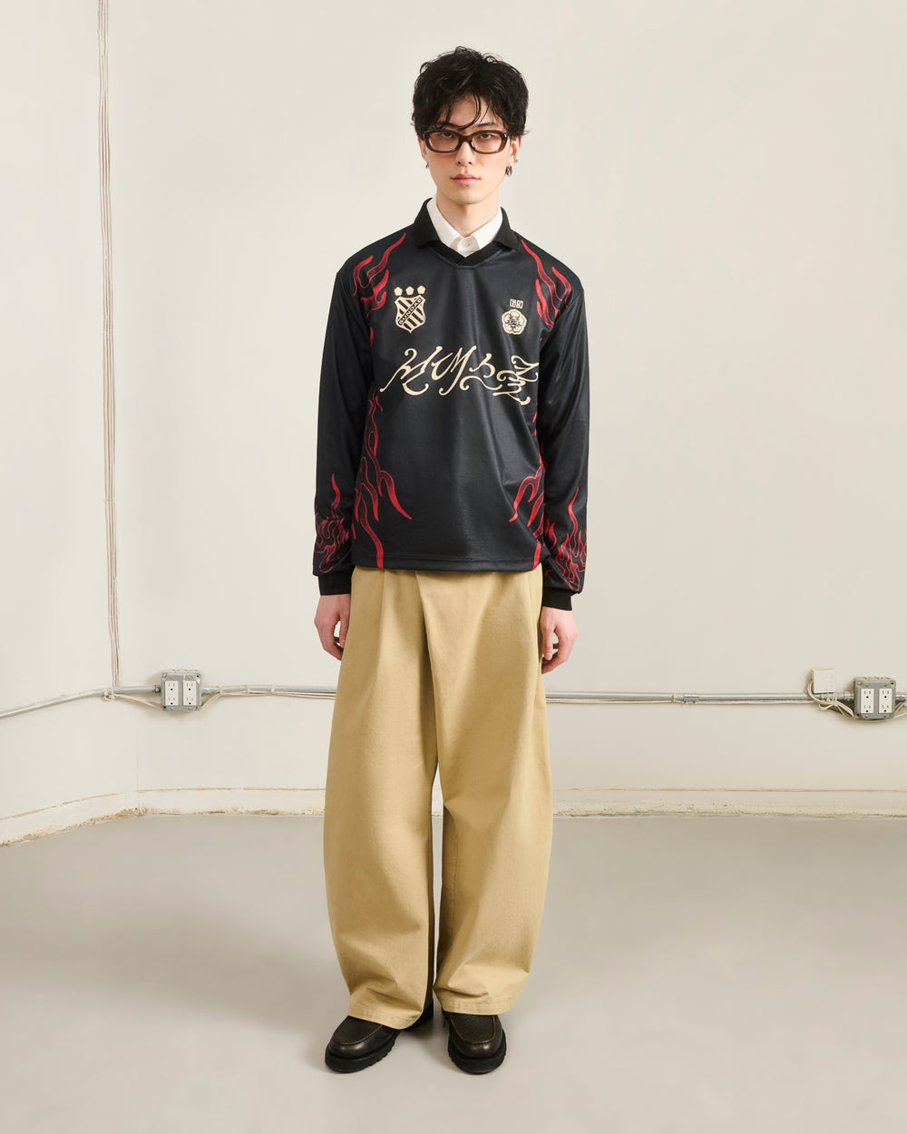 A person with short dark hair and glasses stands against a plain wall, wearing Sundae School's KOREATOWN FOOTBALL JERSEY—featuring custom Korean graphics and red flames—paired with wide beige pants and black shoes, channeling K-Town fashion.