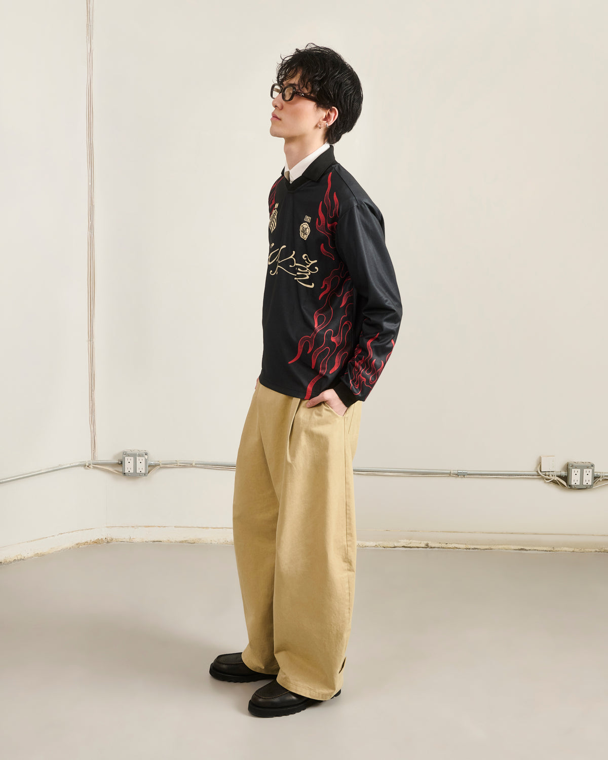 A person stands sideways in a plain room, hands in pockets, wearing Sundae School’s KOREATOWN FOOTBALL JERSEY with red Korean-inspired flame designs, beige wide-leg pants, black shoes, and dark glasses.