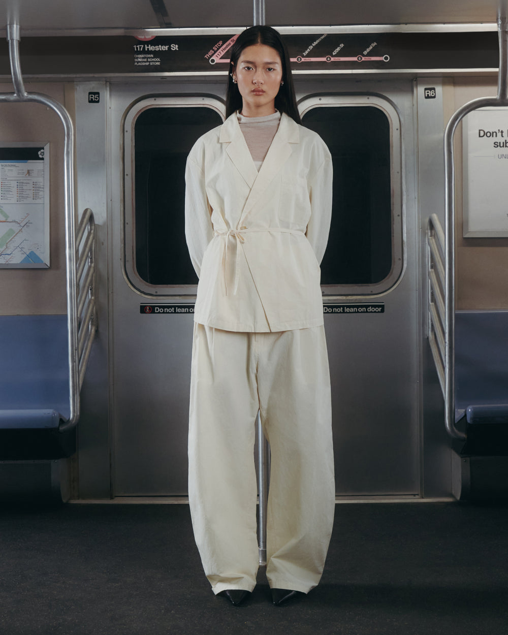 A person wearing Sundae School’s IVORY FLOWER NORIGAE LINEN TROUSERS stands in the middle of an empty subway car, facing forward with their hands behind their back. The subway seats are blue and signs can be seen in the background.