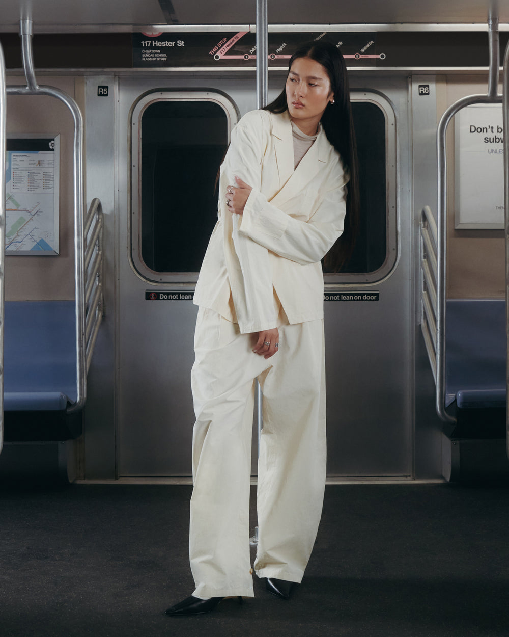 A person wearing Sundae School's IVORY FLOWER NORIGAE LINEN TROUSERS and black shoes stands in a subway car, posing with arms crossed. Subway maps and warning signs are visible on the walls behind them.