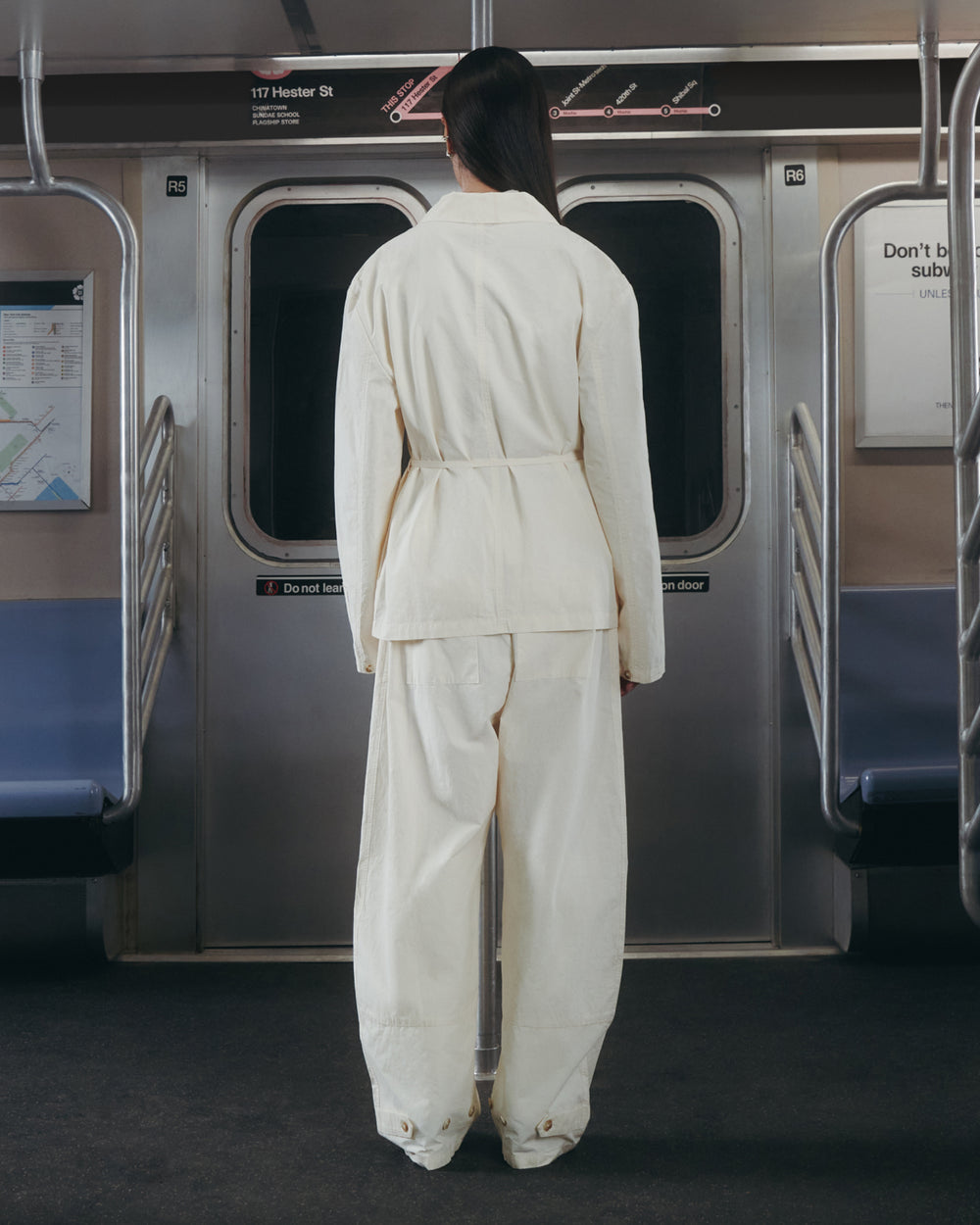 Wearing Sundae School's IVORY FLOWER NORIGAE LINEN TROUSERS, a person in loose, off-white clothing stands with their back to the camera, facing closed subway doors in a car lined with empty blue seats and a map on the left wall.