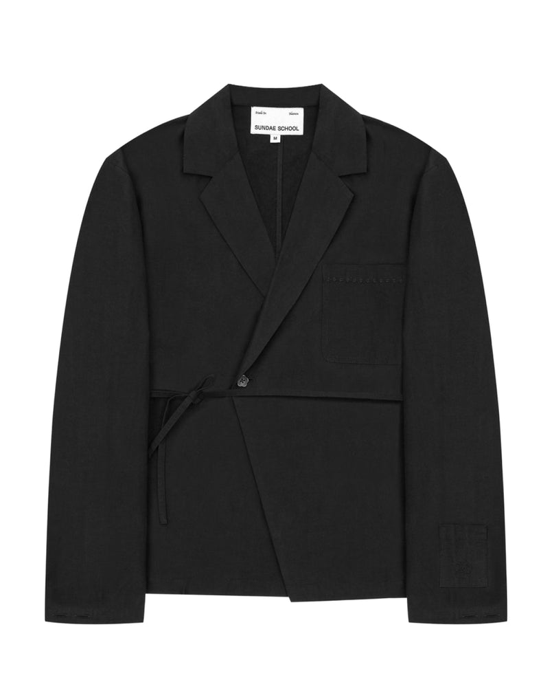The Sundae School BLACK LINEN GORUM BLAZER is an unstructured black wrap-style blazer featuring a single button, left chest pocket, and a tie closure at the waist, displayed against a white background.