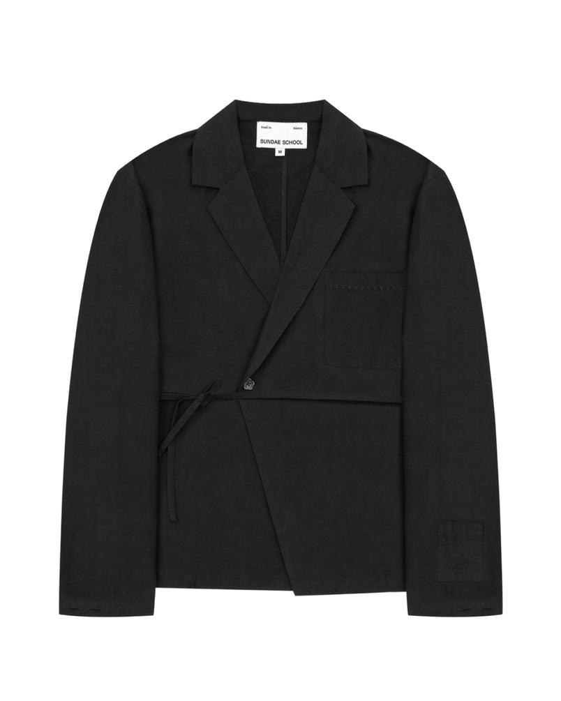 The Sundae School BLACK LINEN GORUM BLAZER is an unstructured black wrap-style blazer featuring a single button, left chest pocket, and a tie closure at the waist, displayed against a white background.