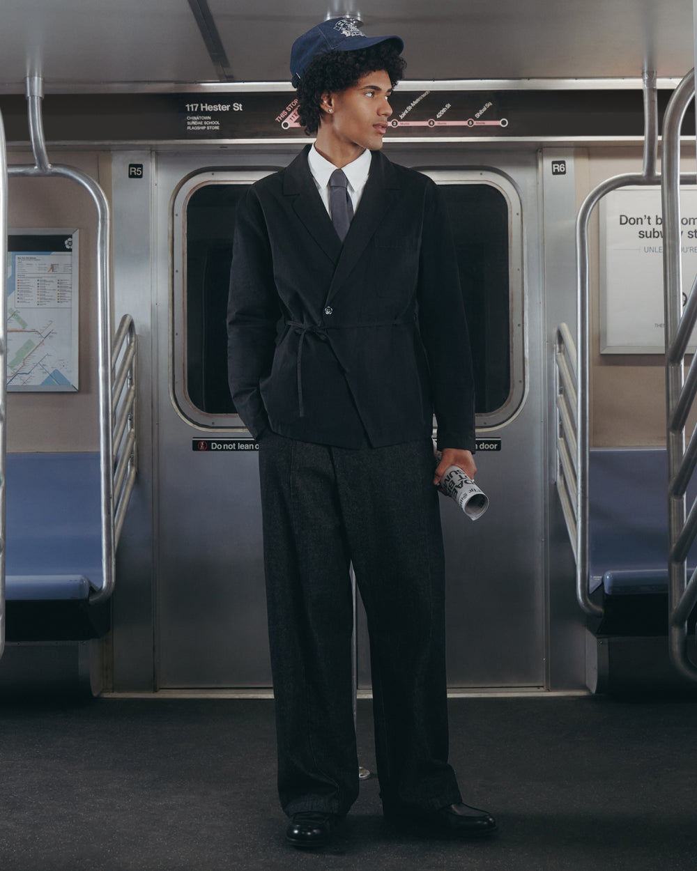 Wearing the BLACK LINEN GORUM BLAZER by Sundae School, a person in a dark suit, tie, and blue bucket hat stands inside a subway car holding a rolled-up newspaper. They look to the side, with empty seats and subway signs visible in the background.