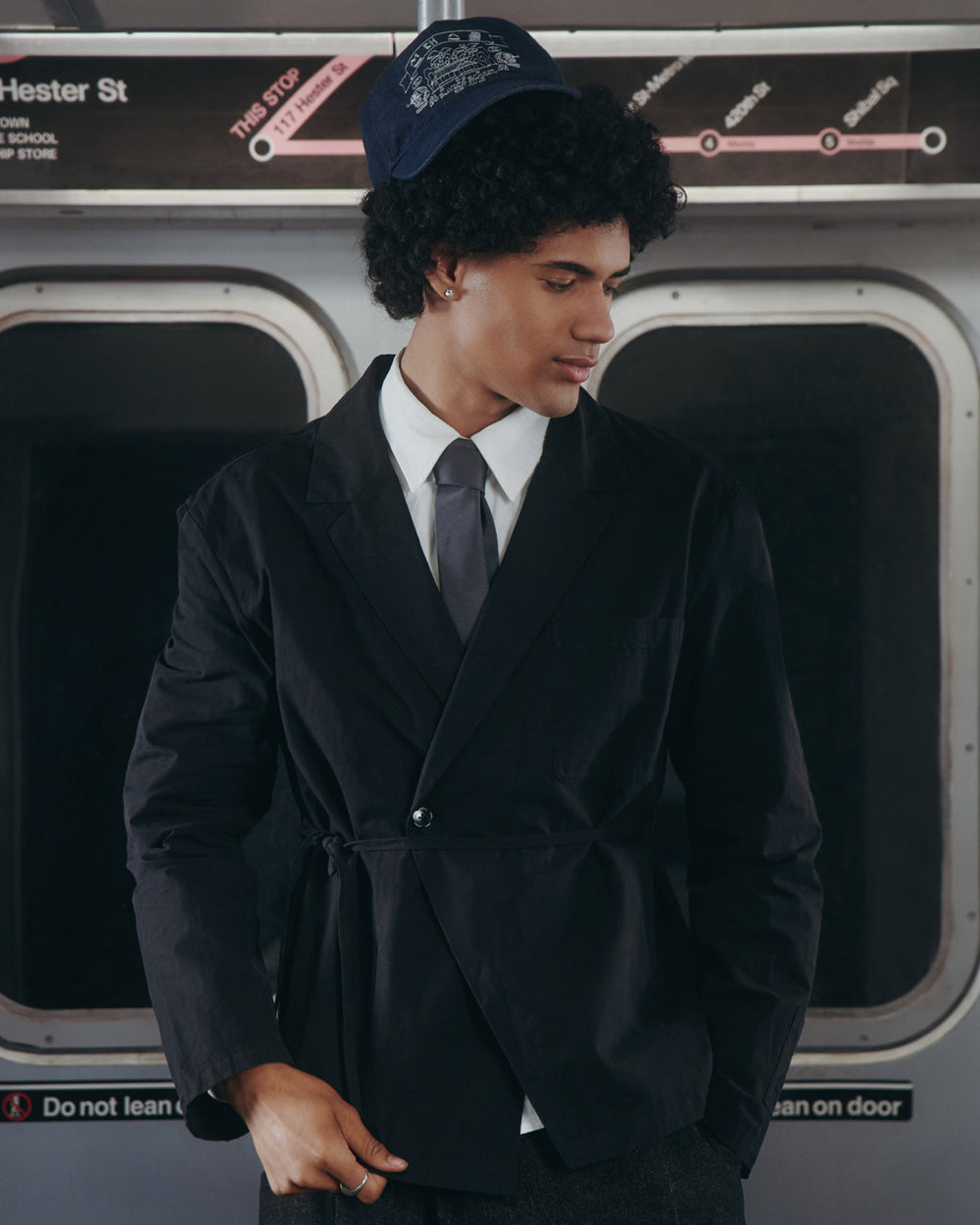 A young man wearing the BLACK LINEN GORUM BLAZER by Sundae School, paired with a white shirt and tie, stands on a subway train. He looks down beneath a navy cap, with “Hester St” and subway signs visible in the background.