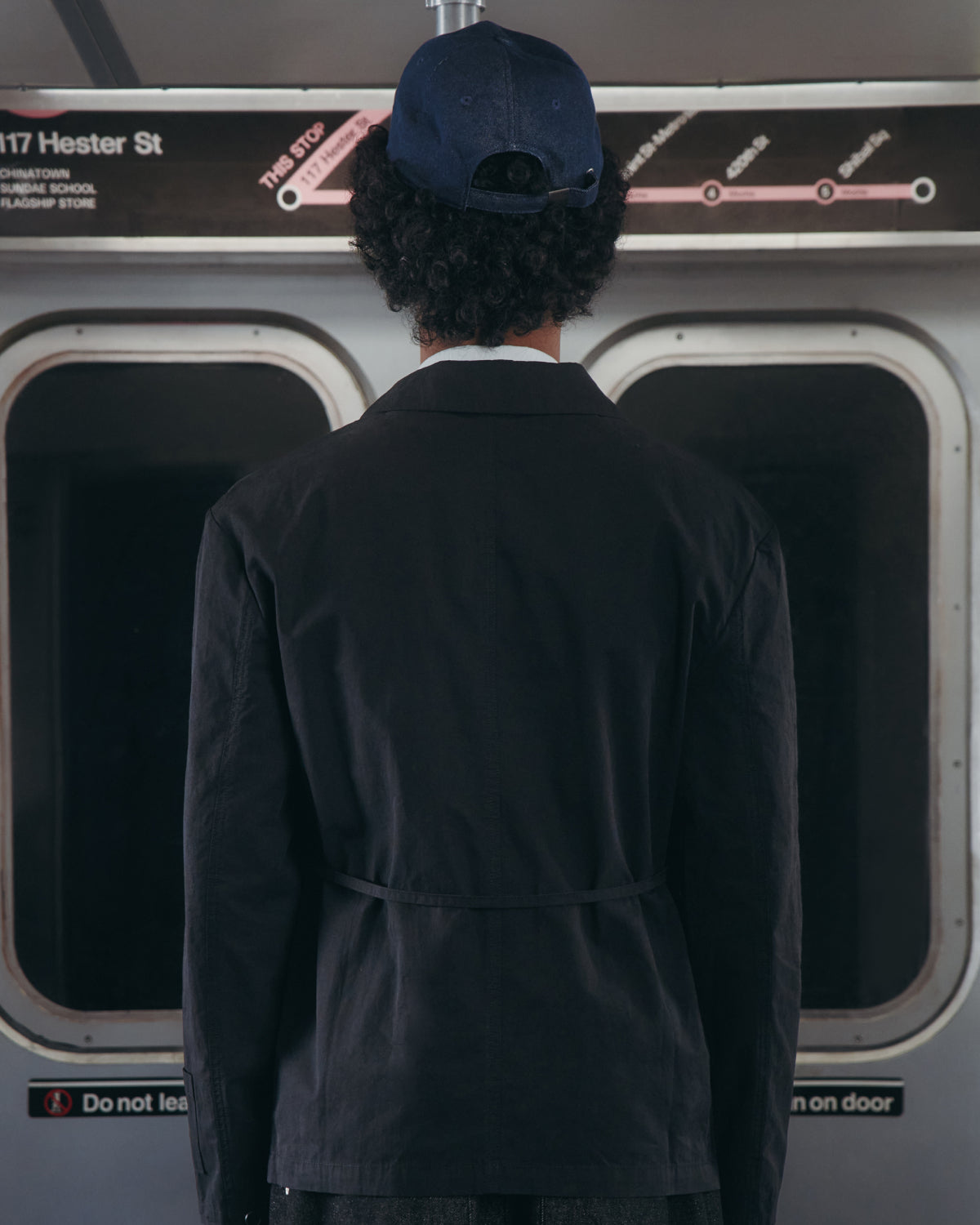 A person wearing the BLACK LINEN GORUM BLAZER from Sundae School and a blue cap stands with their back to the camera inside a subway train, facing the window. The subway map and Hester St station sign are visible above the window.