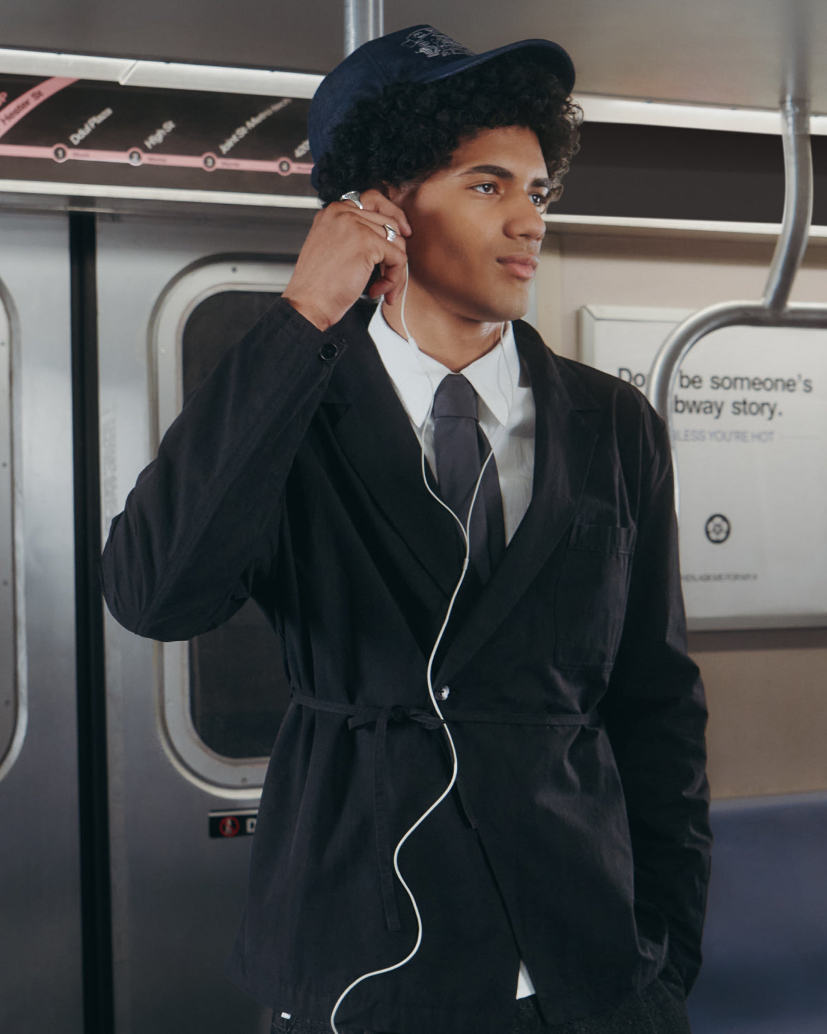 A young man in a BLACK LINEN GORUM BLAZER by Sundae School stands on a subway train, wearing a suit and tie with a blue cap, listening to earphones, one hand adjusting an earbud as he looks to the side.
