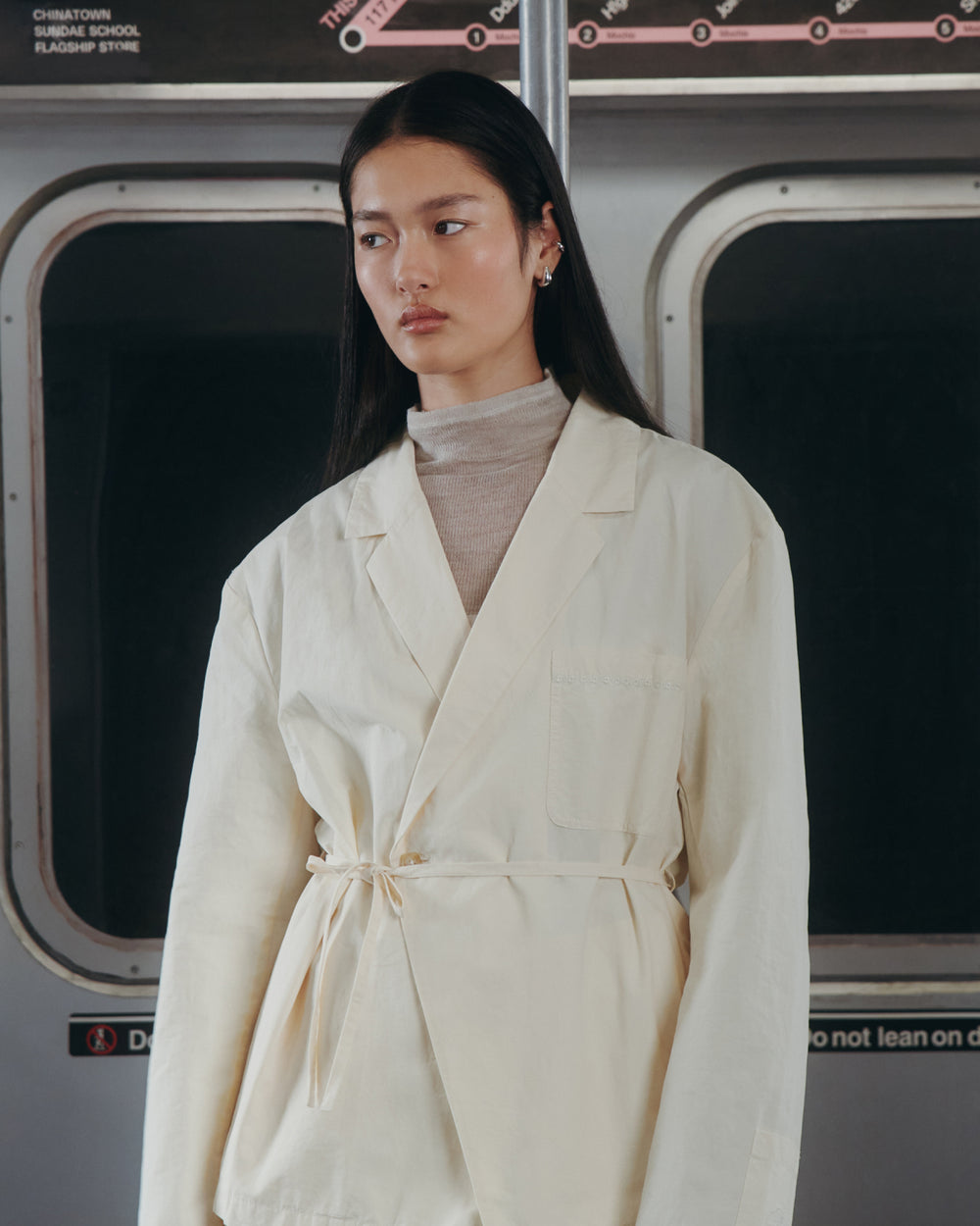 A woman stands in a subway car, wearing the IVORY LINEN GORUM BLAZER by Sundae School over a beige turtleneck. She has long, straight dark hair and a neutral expression.