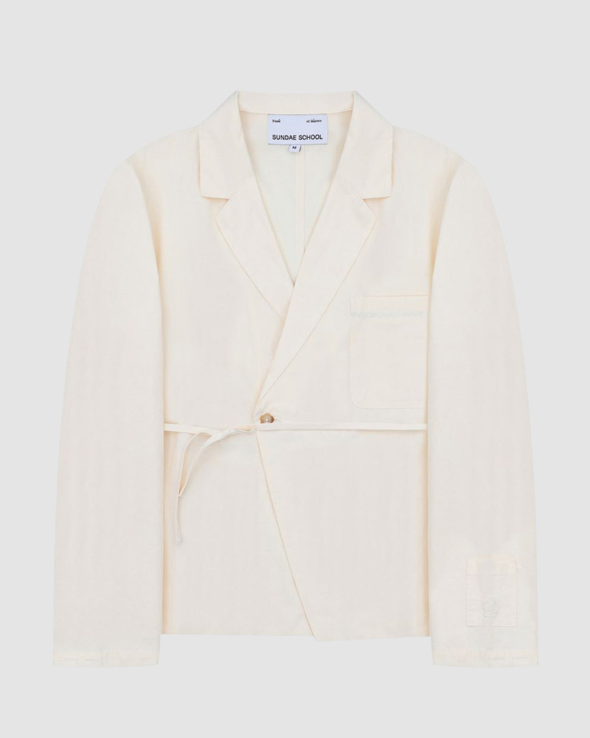 The IVORY LINEN GORUM BLAZER by Sundae School is a cream-colored, single-breasted blazer featuring a notched lapel, front button closure, waist tie, and chest pocket, displayed against a plain light background.