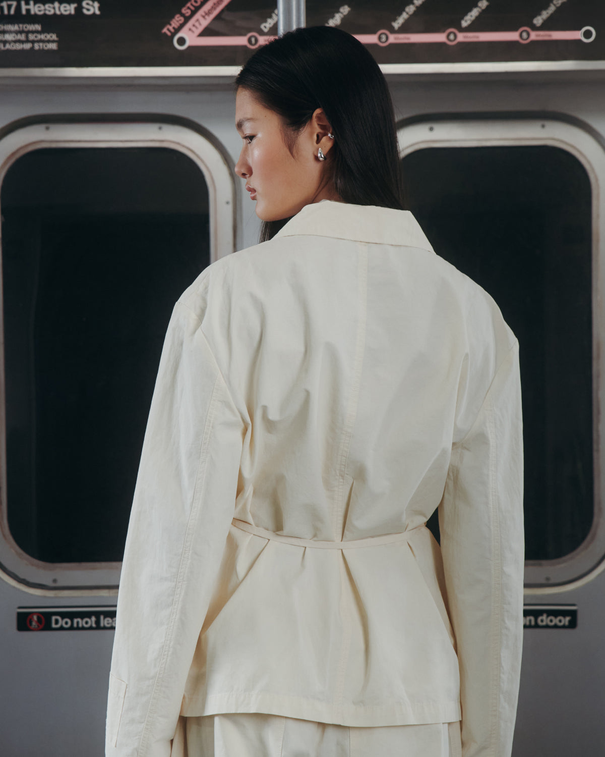 A woman with long dark hair, dressed in the Sundae School IVORY LINEN GORUM BLAZER, stands in front of subway doors, facing away and glancing to the side, with subway maps and signs displayed above.