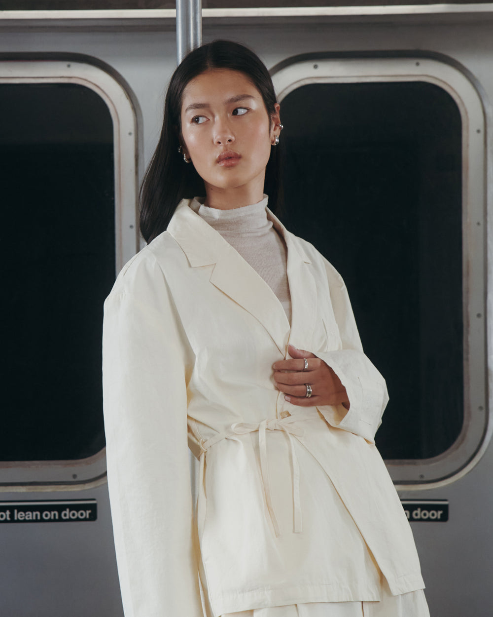 A woman wearing the IVORY LINEN GORUM BLAZER by Sundae School and a turtleneck stands in front of subway doors, gazing calmly to the side.