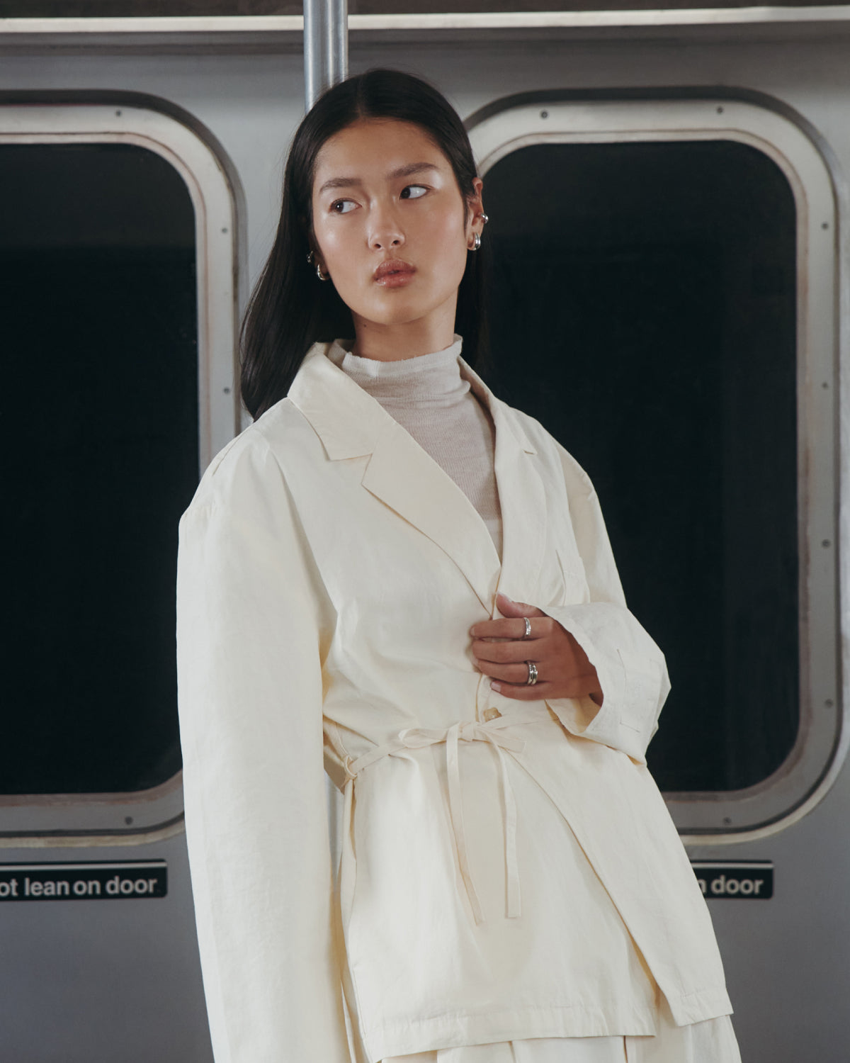 A woman wearing the IVORY LINEN GORUM BLAZER by Sundae School and a turtleneck stands in front of subway doors, gazing calmly to the side.