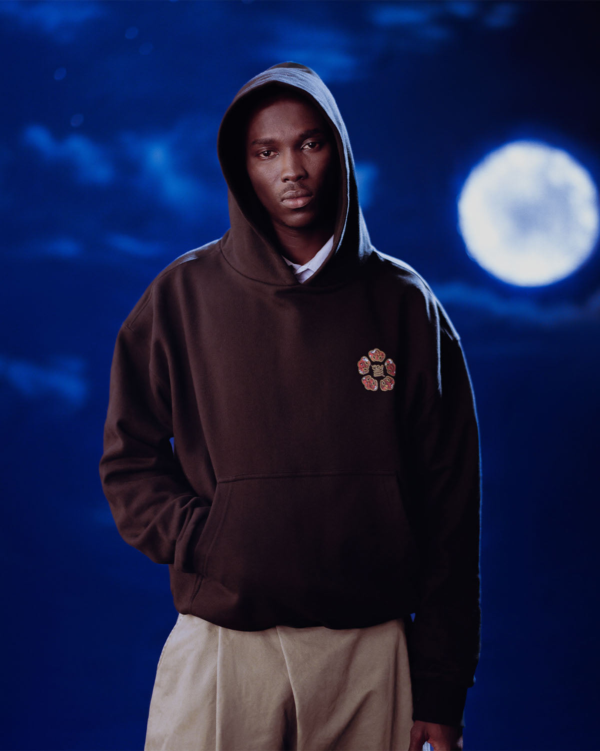 Someone wears the HIGH HORSE LNY HOODIE by Sundae School with light pants, standing with one hand in their pocket against a night sky featuring a full moon and clouds.