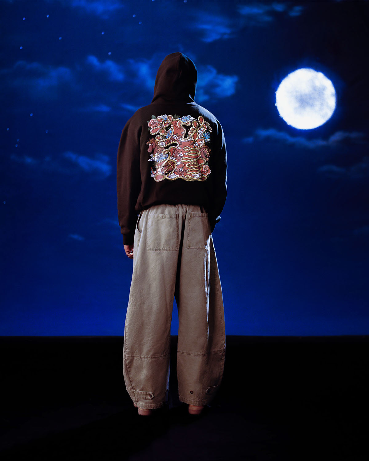 A person wearing the Sundae School HIGH HORSE LNY HOODIE with a colorful back design and wide beige pants stands facing a night sky illuminated by a full moon and scattered clouds.