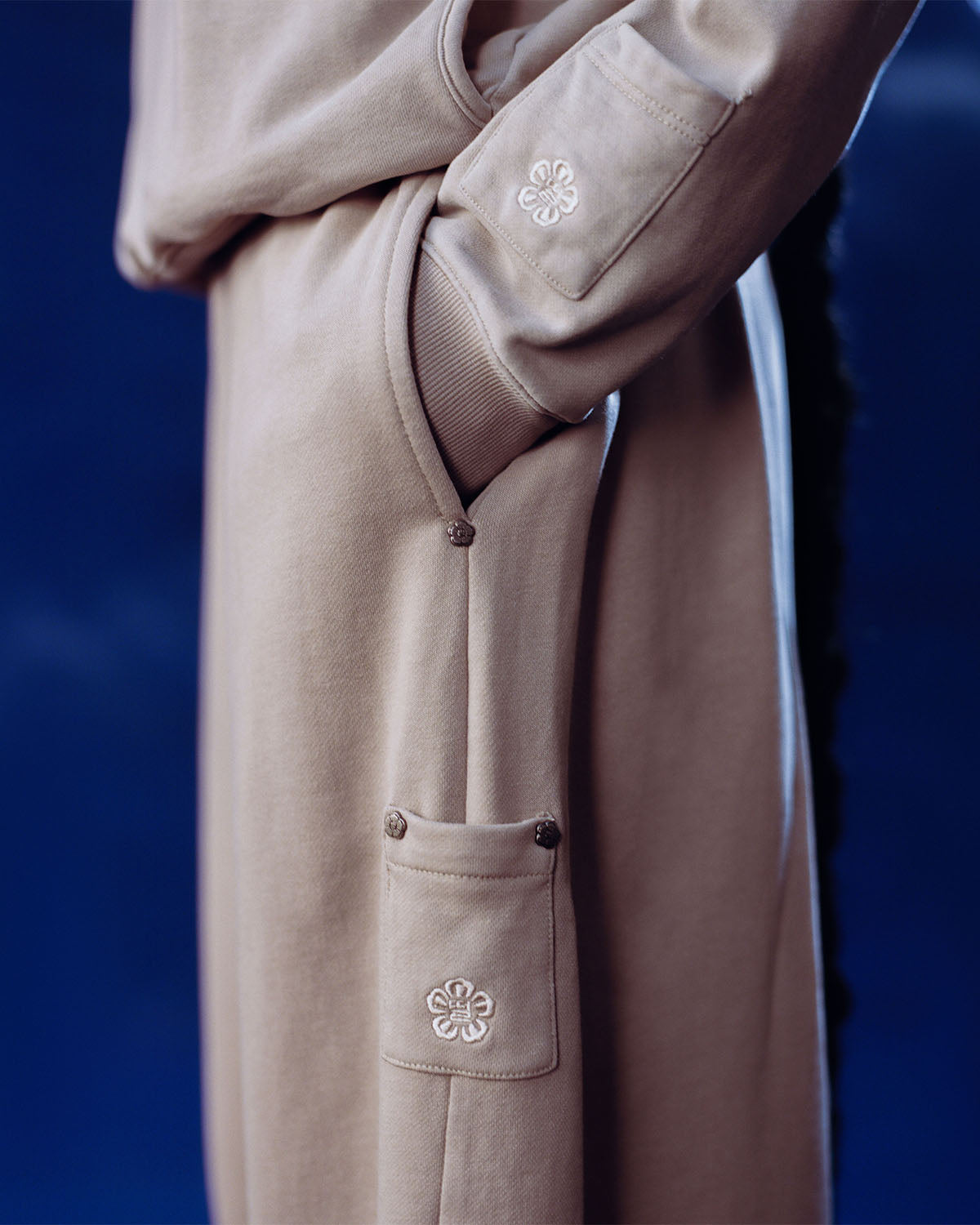 A person wears Sundae School's TAUPE WRAP SWEATBAJI, featuring embroidered white florals and metal snap buttons on the sleeve and side pocket. Made from 100% cotton, the beige Hanbok-inspired pants are shown against a dark blue background.
