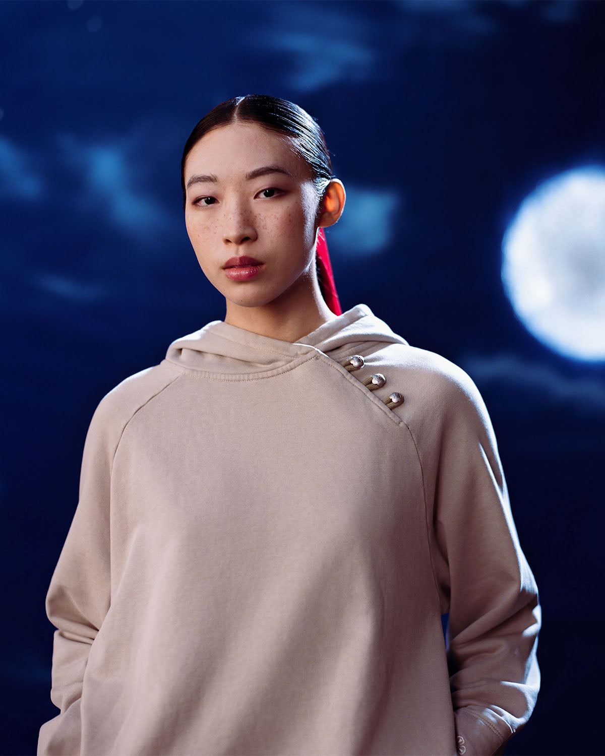 A person with long dark hair tied back wears the Sundae School TAUPE HANBOK HOODIE, featuring a custom “떨” logo, asymmetric hood closure, and signature stash pocket, standing before a night sky with a glowing full moon.
