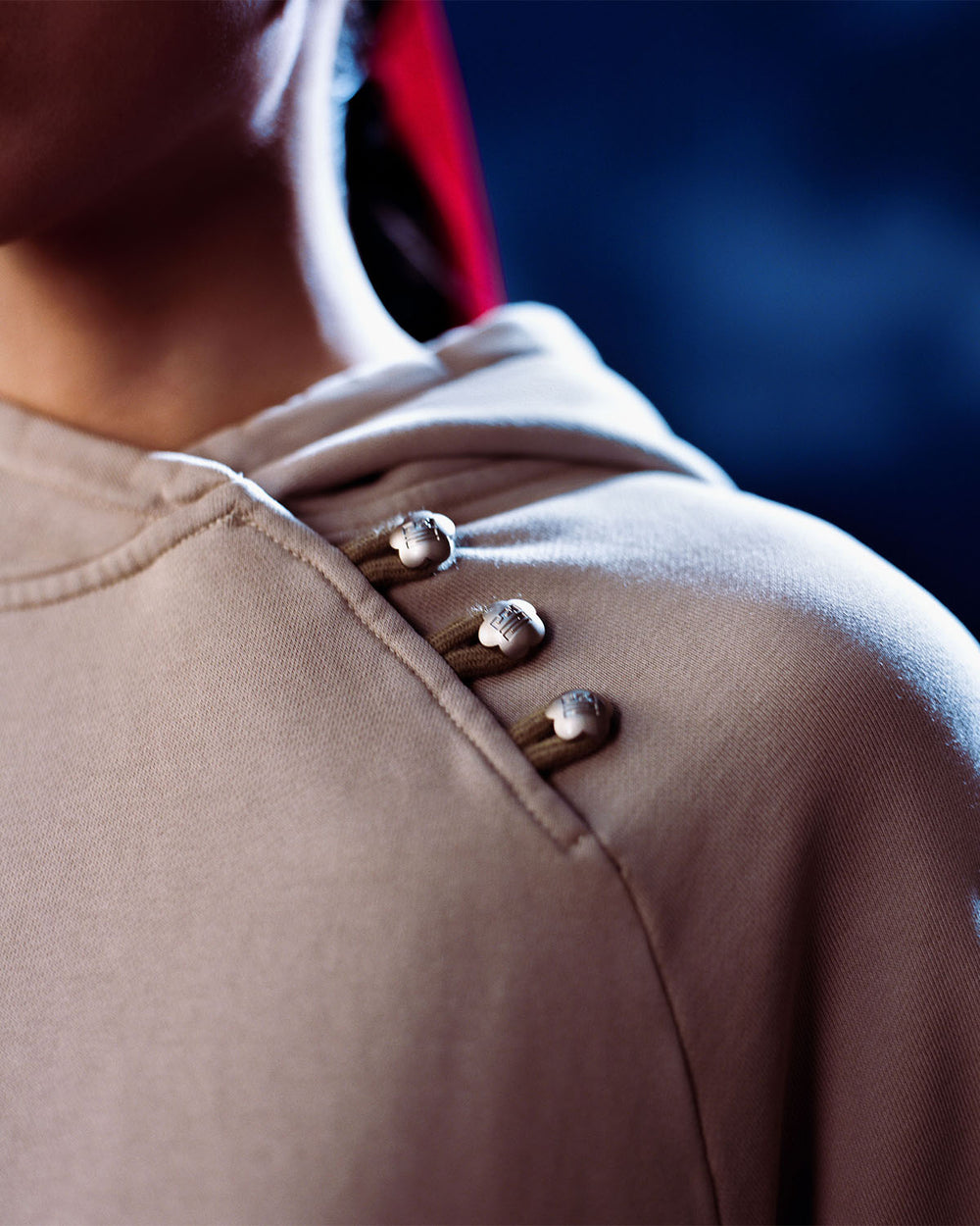 A close-up of a person wearing the Sundae School TAUPE HANBOK HOODIE, featuring an asymmetric hood closure and three silver domed buttons fastened with loops on the shoulder. The background is blurred in dark blue tones.