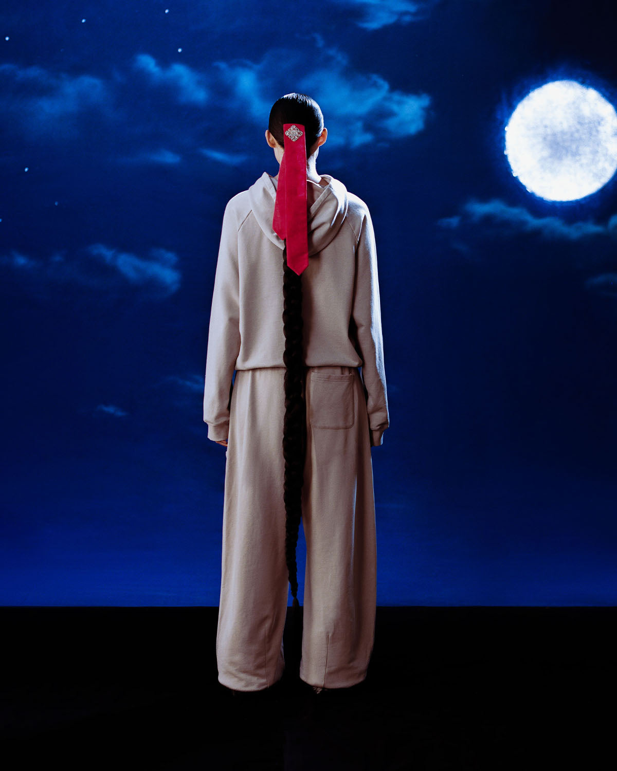 Dressed in Sundae School's TAUPE WRAP SWEATBAJI, a person faces away from the camera under a bright full moon, their long braid tied with a red ribbon, against a night sky scattered with clouds.
