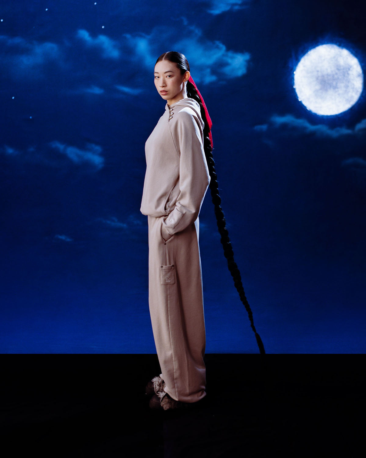 A woman with a long braided ponytail and red ribbon poses in Sundae School’s TAUPE WRAP SWEATBAJI, Hanbok-inspired loungewear pants, against a night sky backdrop with clouds and a full moon.