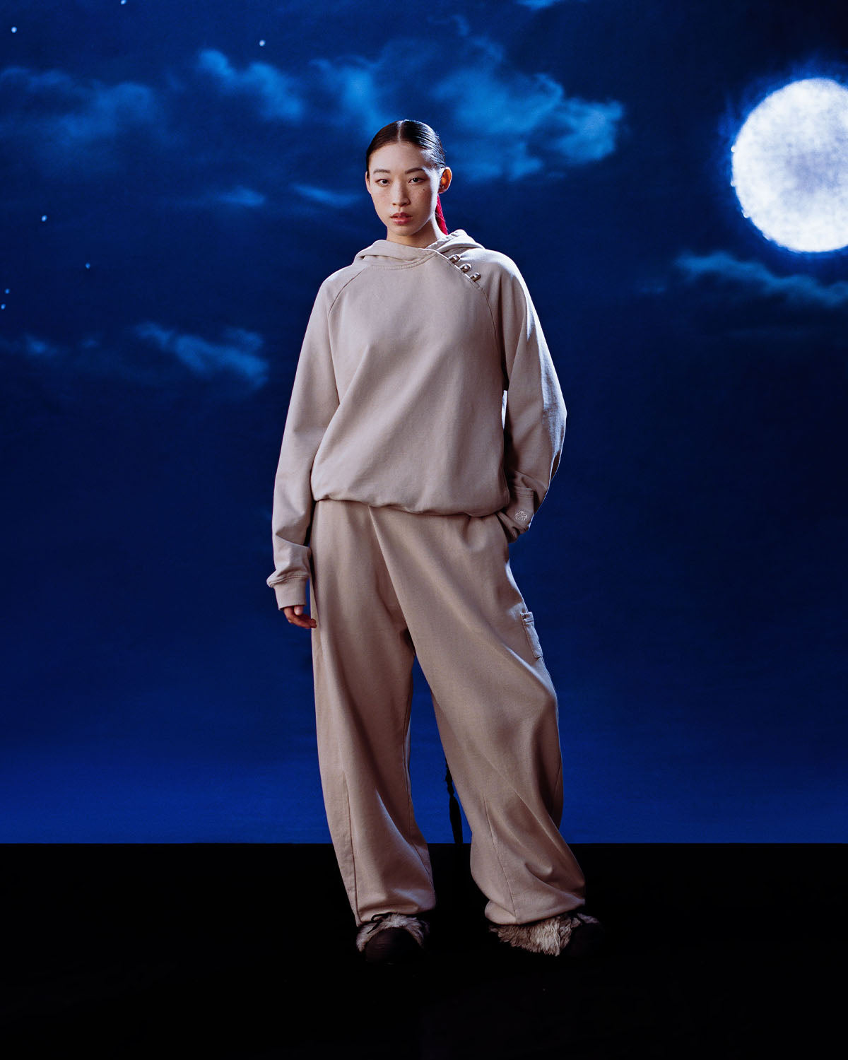 A person wears Sundae School's TAUPE WRAP SWEATBAJI, Hanbok-inspired pants, and a loose beige hoodie, standing against a dark blue night sky with a full moon and scattered stars.