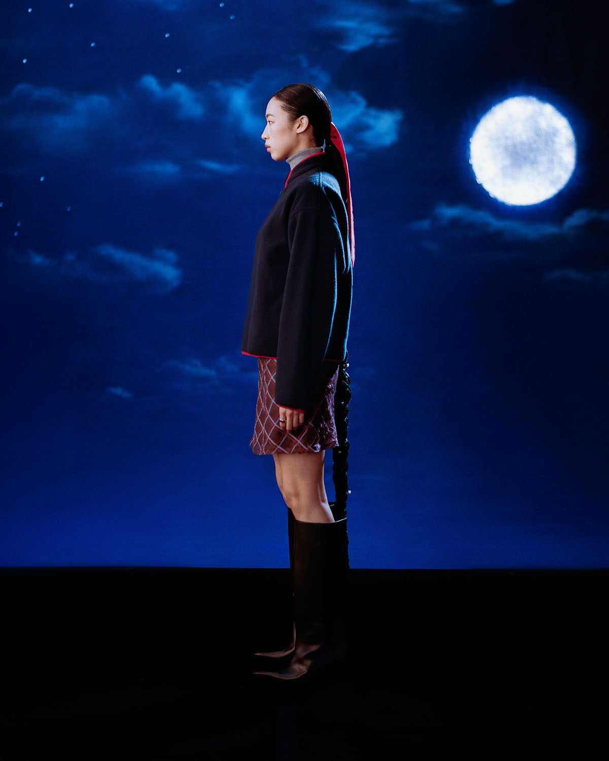 A woman in profile stands against a dark blue night sky with a full moon and scattered clouds, wearing the Sundae School NAVY TEARDROP KNOT FLEECE, a patterned skirt, tall boots, and her hair in a long ponytail.
