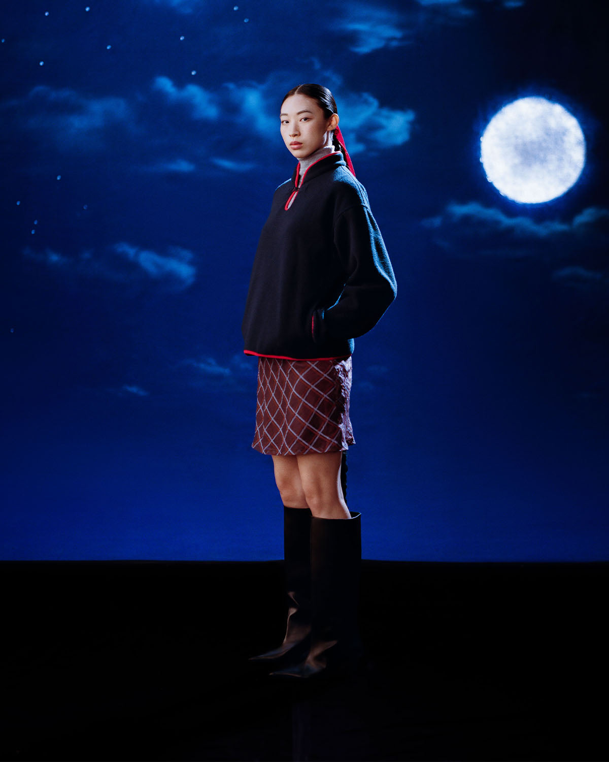 A woman stands confidently before a moonlit sky in the Sundae School NAVY TEARDROP KNOT FLEECE, paired with a patterned skirt and tall black boots, her hair pulled back in a ponytail.