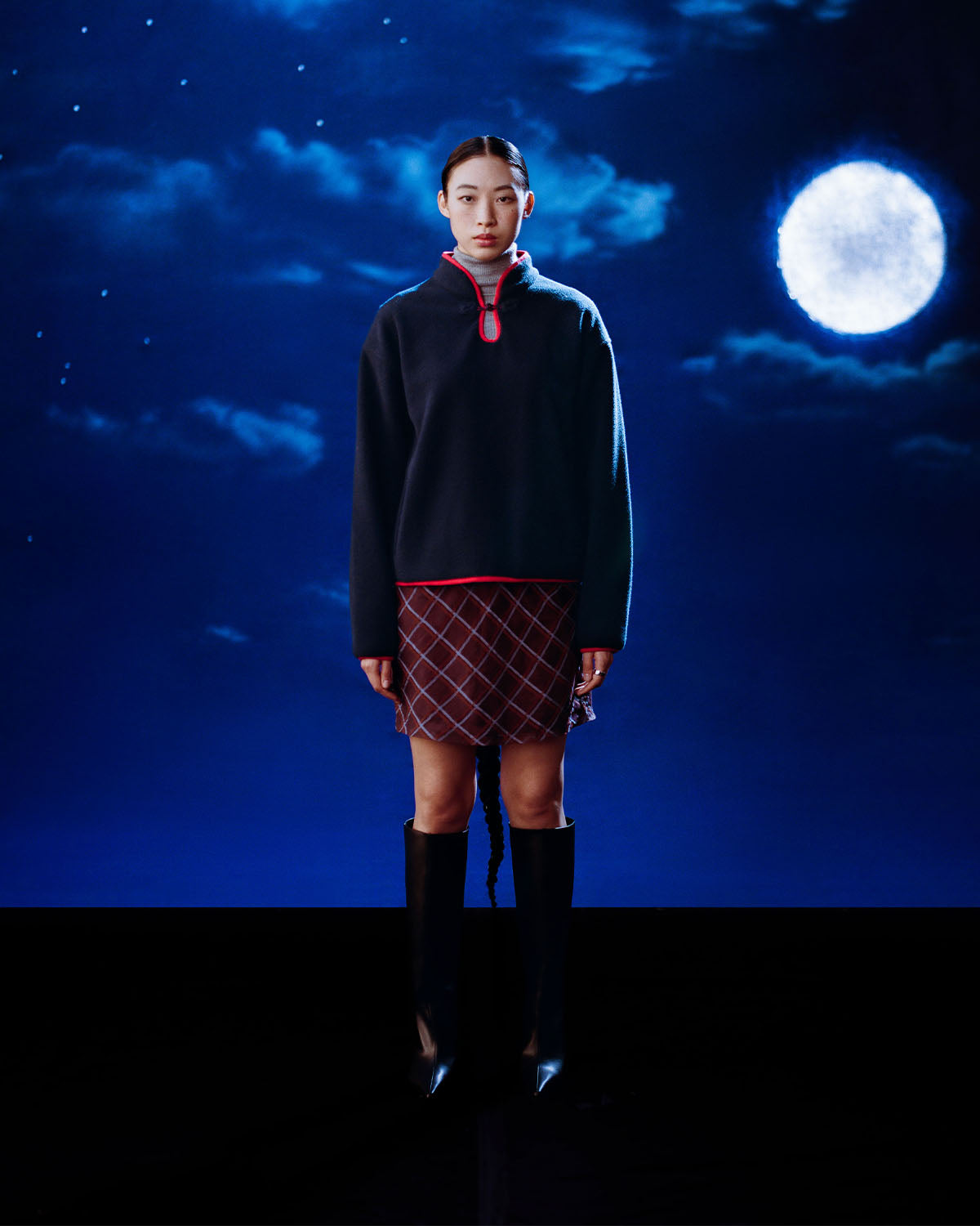 A woman faces the camera against a dark, moonlit sky, wearing Sundae School's NAVY TEARDROP KNOT FLEECE sweater with red trim, a patterned skirt, and knee-high black boots, her expression neutral.