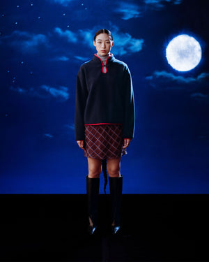 A woman faces the camera against a dark, moonlit sky, wearing Sundae School's NAVY TEARDROP KNOT FLEECE sweater with red trim, a patterned skirt, and knee-high black boots, her expression neutral.