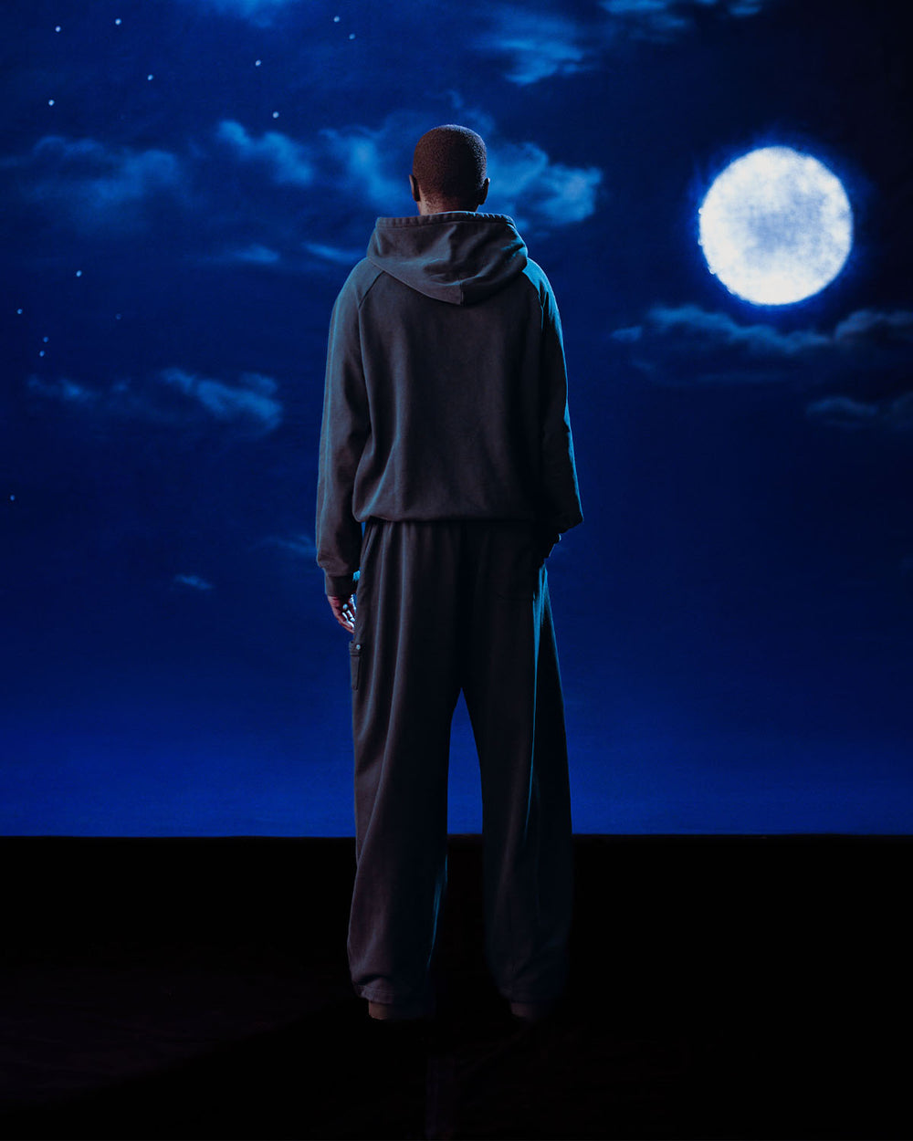 A man stands before a moon, wearing Sundae School’s INDIGO WASH HANBOK HOODIE featuring a custom "떨" logo and signature sleeve stash pocket.