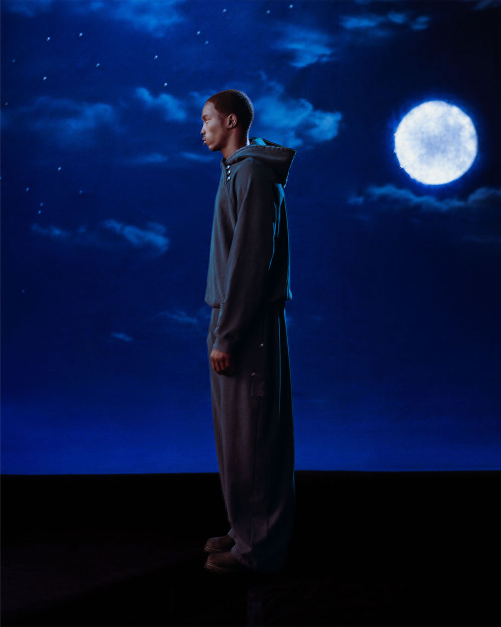 A person in loose, dark clothing stands in profile against a night sky, lit by a full moon, wearing Sundae School's INDIGO WASH HANBOK HOODIE featuring a custom "떨" logo and signature sleeve stash pocket.