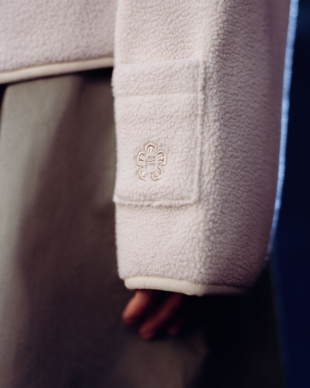 A close-up of a person's arm in Sundae School's TAUPE TEARDROP KNOT FLEECE, a cream-colored East Asian-inspired jacket with an embroidered pocket, paired with taupe pants and a partially visible hand.