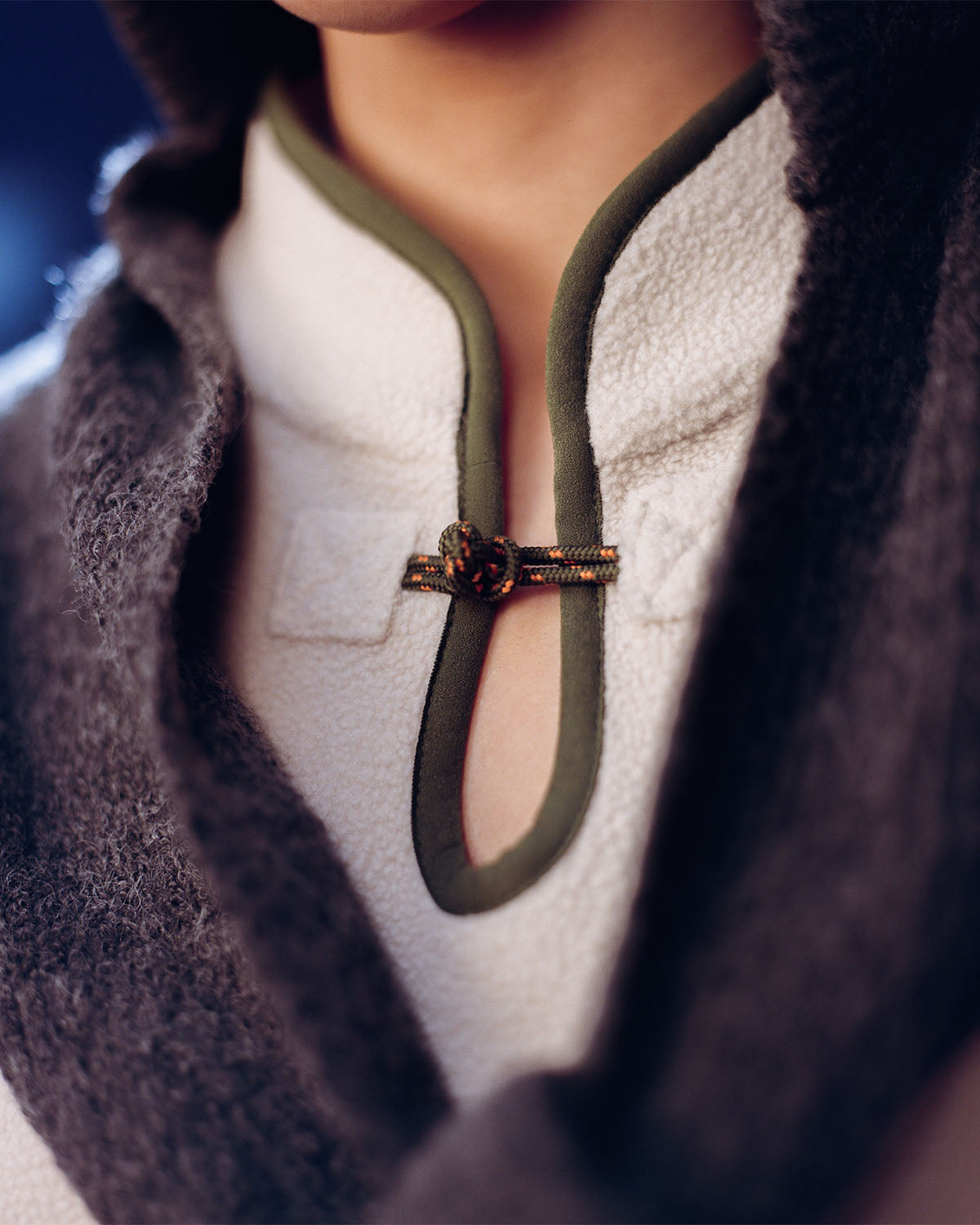 Close-up of a person in Sundae School’s TAUPE TEARDROP KNOT FLEECE, featuring textured white fleece with green trim and an ornate East Asian-inspired clasp at the neckline, partially covered by a gray shawl. Face is partially visible, highlighting cozy details.