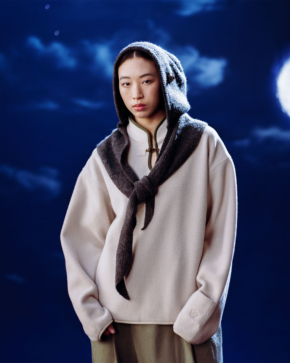 A person stands against a cloudy night sky and bright moon, wrapped in the Sundae School TAUPE TEARDROP KNOT FLEECE, with a gray knitted hood and matching scarf draped over their shoulders.