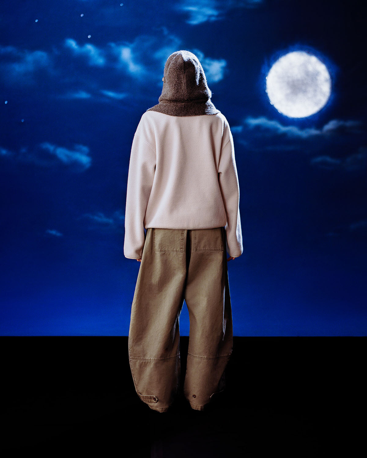 Dressed in the Sundae School TAUPE TEARDROP KNOT FLEECE, a person stands in cozy style facing a dark blue night sky with clouds and a bright full moon.