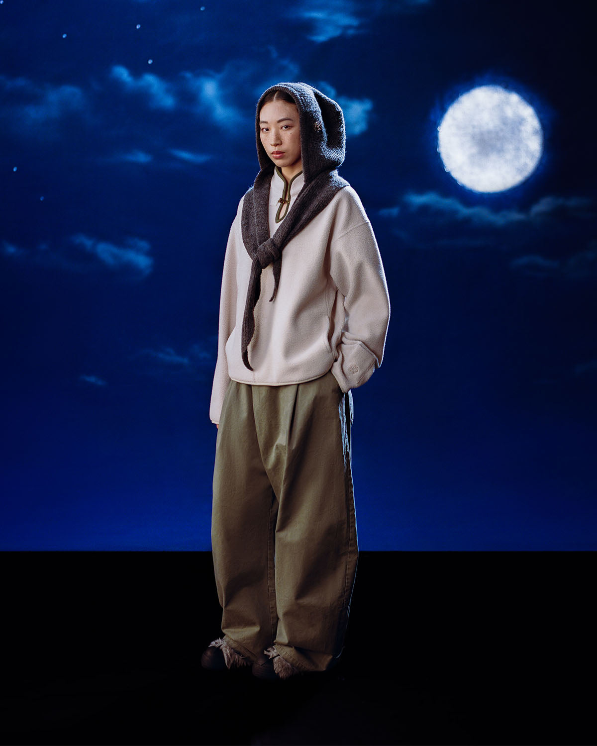 A person in loose, casual clothing stands under a cloudy night sky with a full moon, thoughtfully looking at the camera while wearing the Sundae School TAUPE TEARDROP KNOT FLEECE hat and scarf.