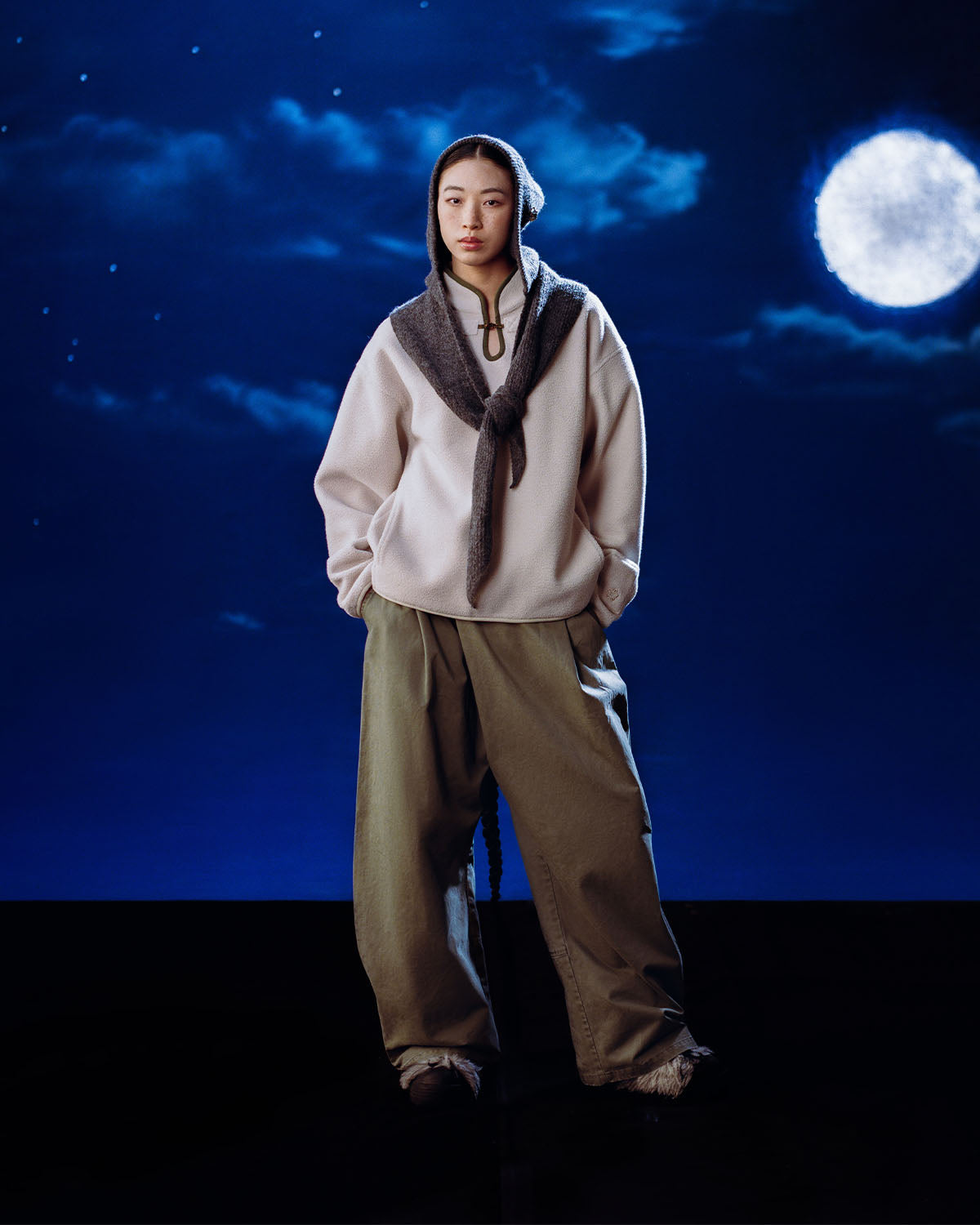 A person stands under a cloudy night sky and full moon, wearing Sundae School’s TAUPE TEARDROP KNOT FLEECE with a furry hood scarf and loose olive-green pants.