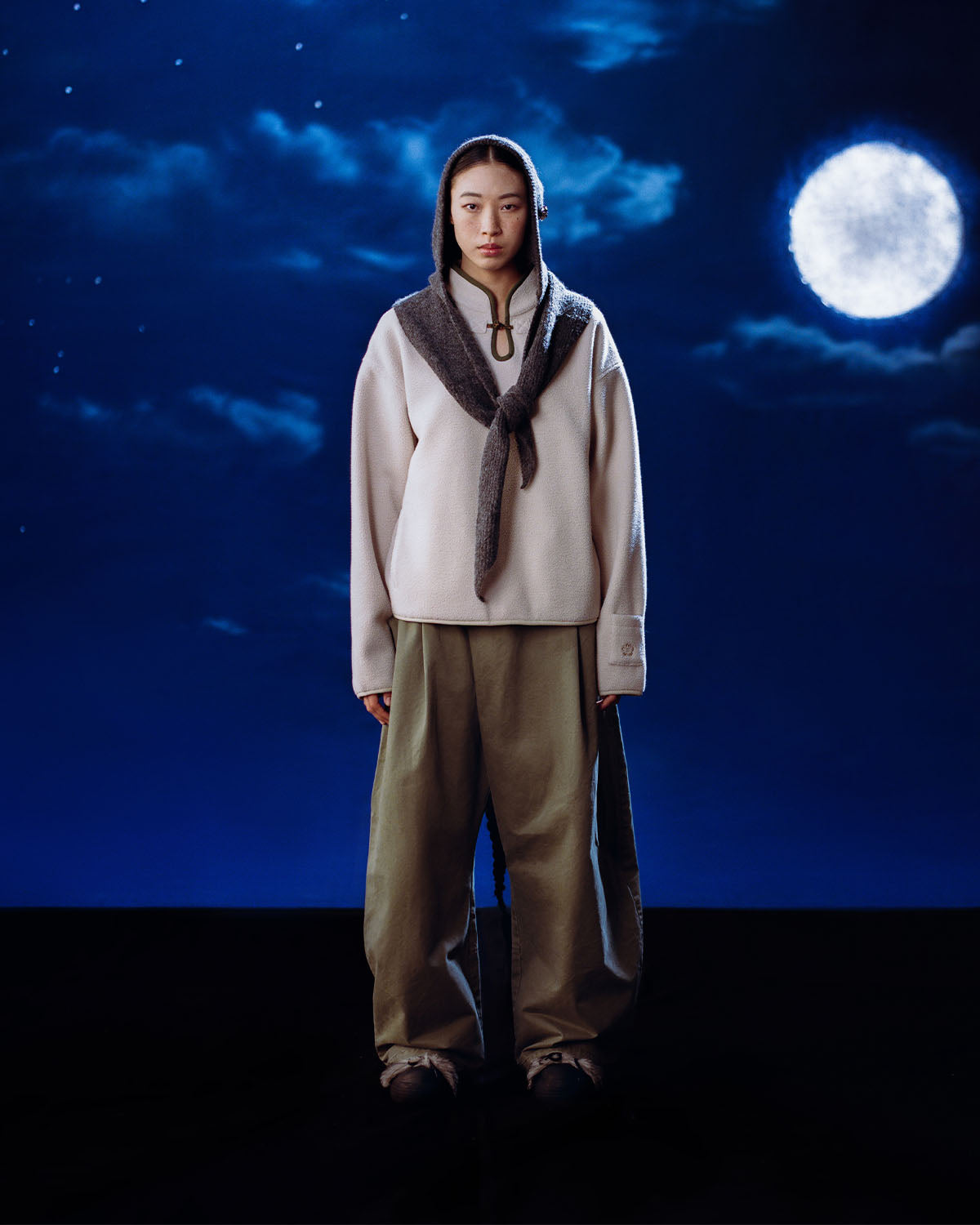 A person stands under a full moon in the night sky, wearing the Sundae School TAUPE TEARDROP KNOT FLEECE with a gray sweater over their shoulders, loose khaki pants, and dark shoes.