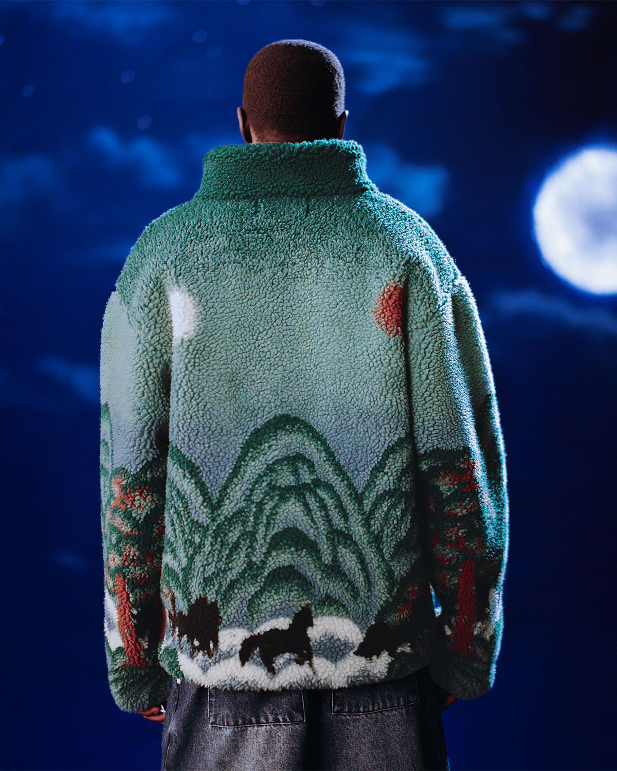 A person faces away wearing Sundae School's TEAL FIVE PEAKS FLEECE, a fuzzy green jacket with a landscape of hills, trees, and horses under a cloudy night sky and bright full moon.