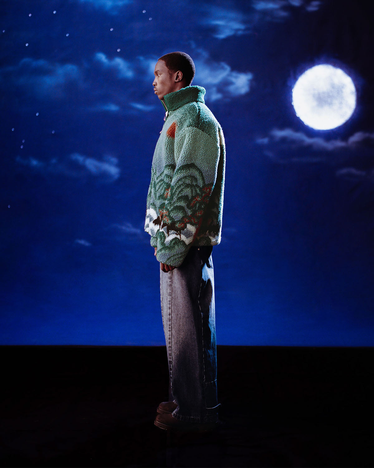 A person stands in profile wearing the Sundae School TEAL FIVE PEAKS FLEECE and wide-leg jeans, set against a nighttime backdrop with a full moon and stars.