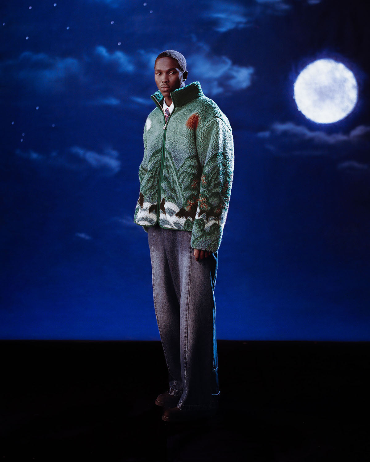 A man wearing the Sundae School TEAL FIVE PEAKS FLEECE stands against a dark, starry night sky with clouds and a bright full moon, paired with wide-leg jeans.