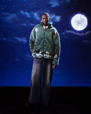 Under a dreamy, dark blue night sky with clouds and a full moon, a person wears Sundae School’s TEAL FIVE PEAKS FLEECE—green with nature patterns—and wide-legged jeans. The surreal atmosphere enhances the look.