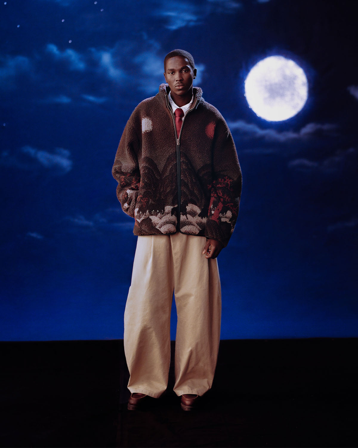 A person in Sundae School's CHARCOAL FIVE PEAKS FLEECE and wide beige pants stands before a nighttime sky with clouds and a full moon.