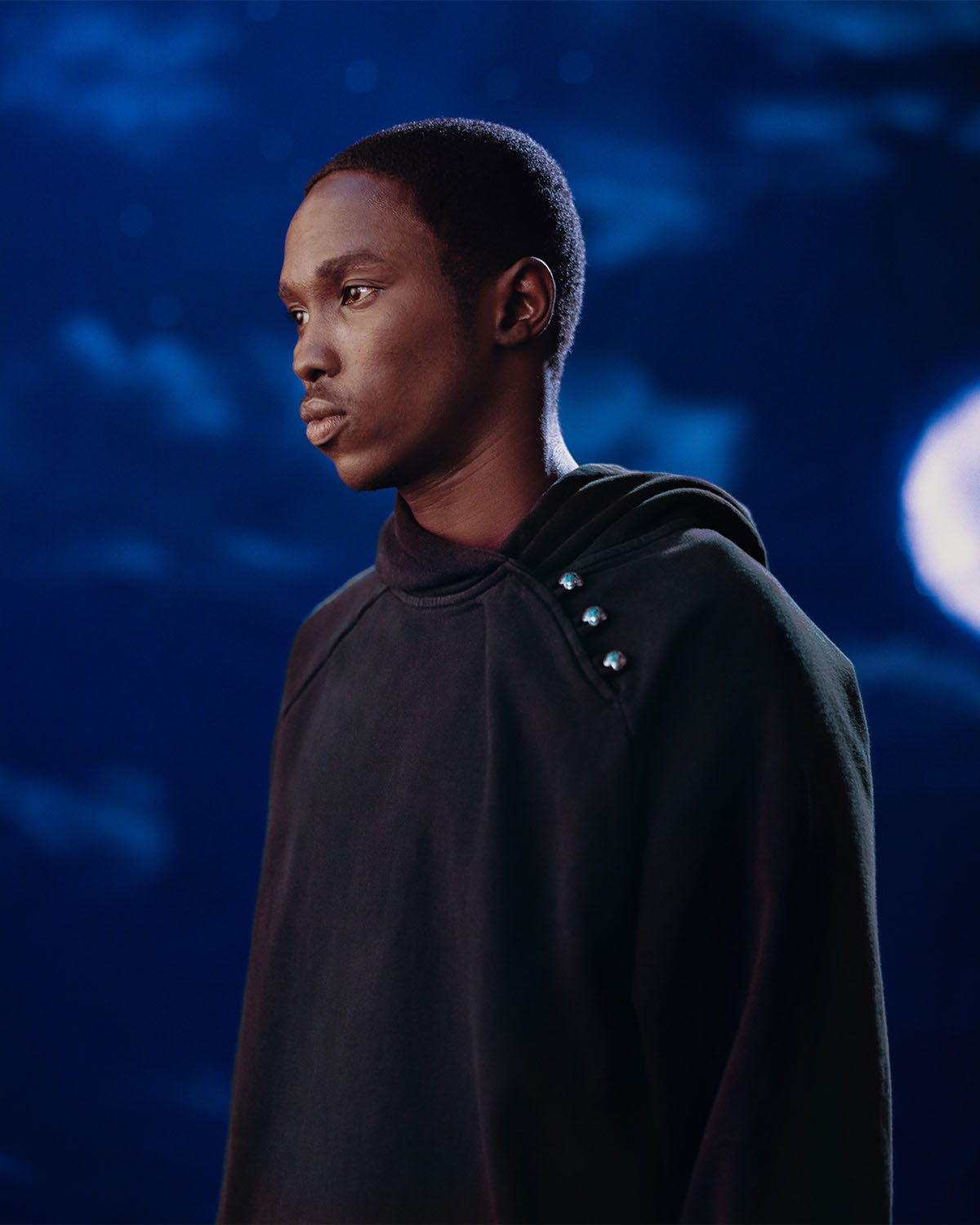 A person in the Sundae School FADE BLACK HANBOK HOODIE, featuring an asymmetric hood closure and button details, stands before a deep blue, outer space-inspired background, gazing thoughtfully to the side.